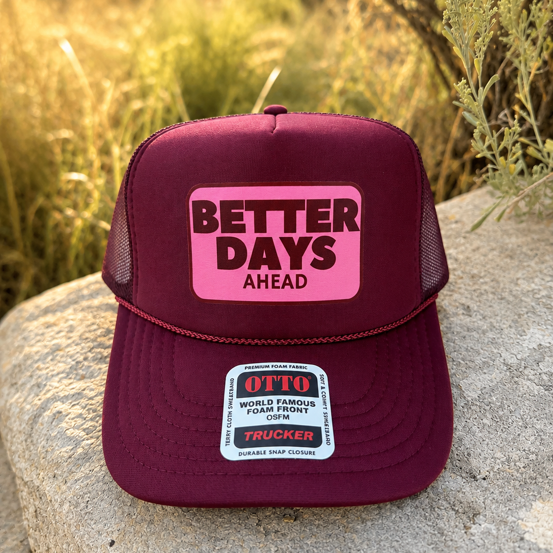 A maroon trucker hat with a pink front patch reading 'BETTER DAYS AHEAD' sits on a large rock outdoors, with dry grass and warm sunlight in the background.